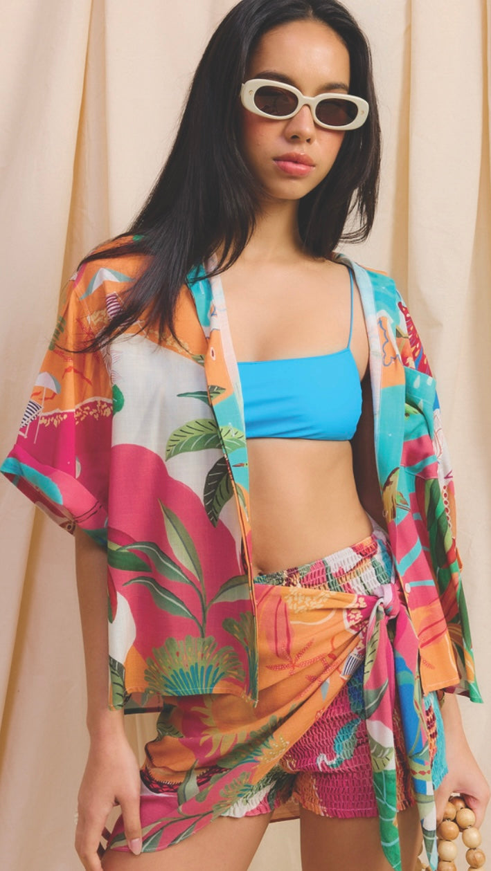 Woman wearing a colorful kimono-style jacket over a blue bikini top and pink shorts against a beige curtain background.